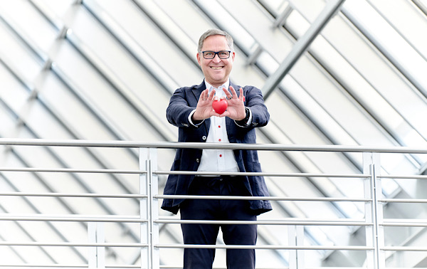 Mann mit Brille und Anzug (Martin) steht oben auf einer Galerie und zeigt ein rotes Herz. mit beiden Händen